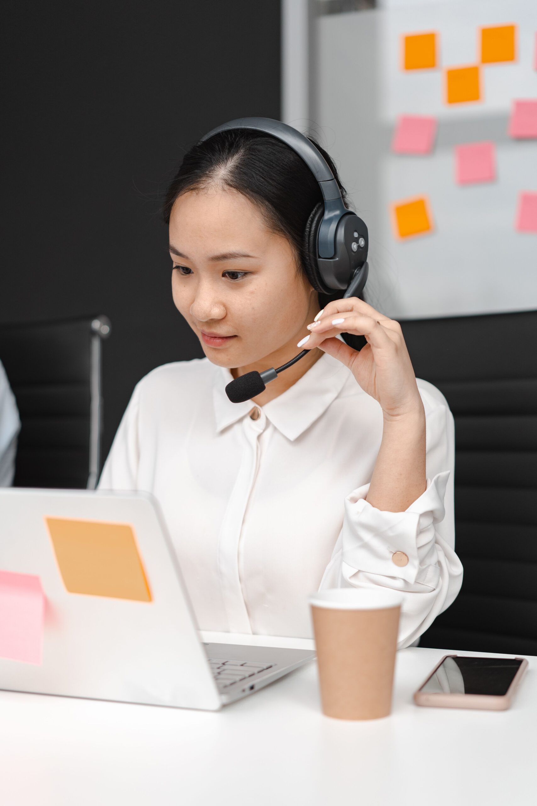 Asian woman using a laptop and headset for customer support in an office environment.